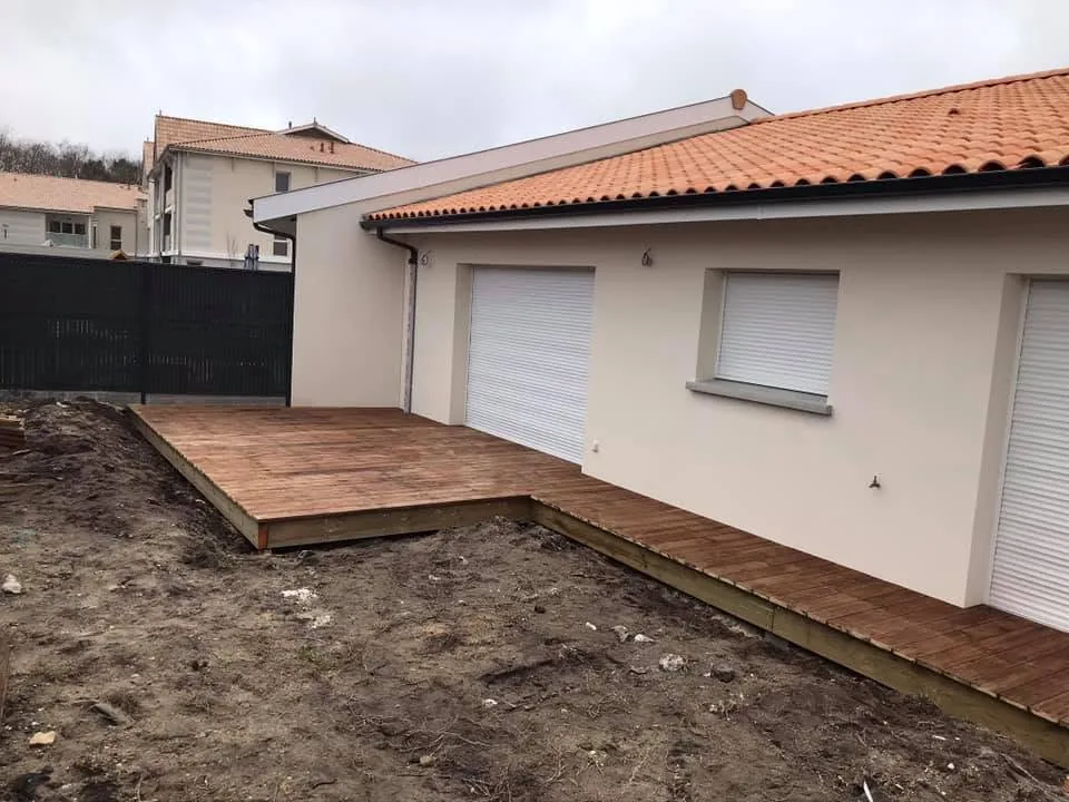 Terrasse en bois devant une maison claire, prolongée par un passage le long de la façade.