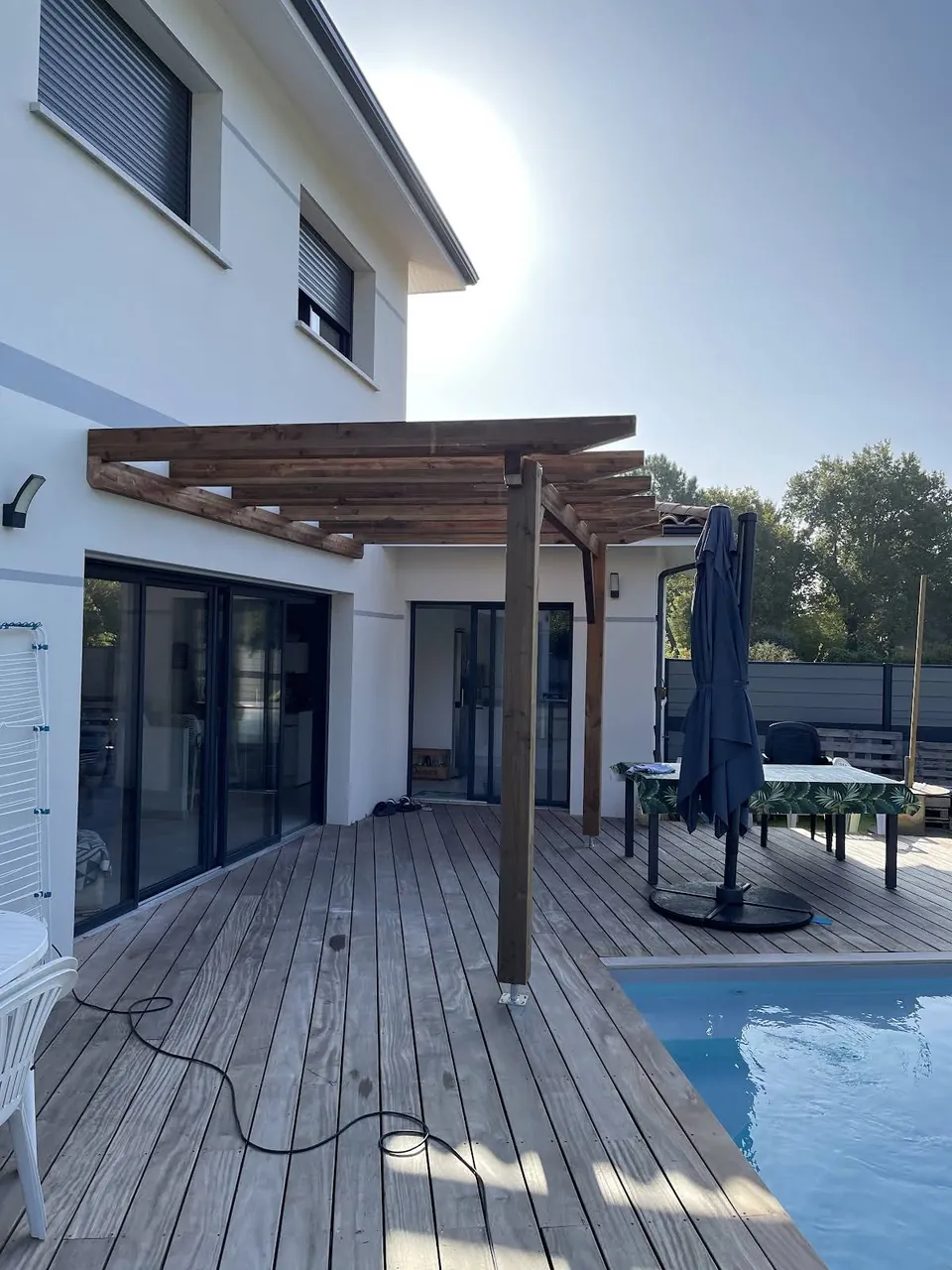 Pergola en bois sur une terrasse au bord d’une piscine, contre une maison blanche.