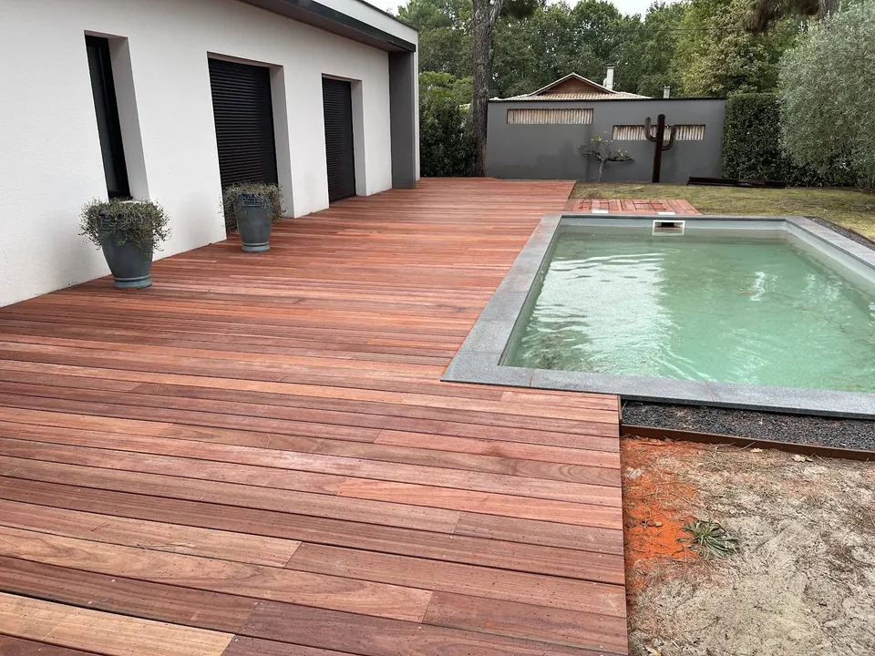 Terrasse en bois aux tons rouges le long d’une piscine et d’une maison blanche.